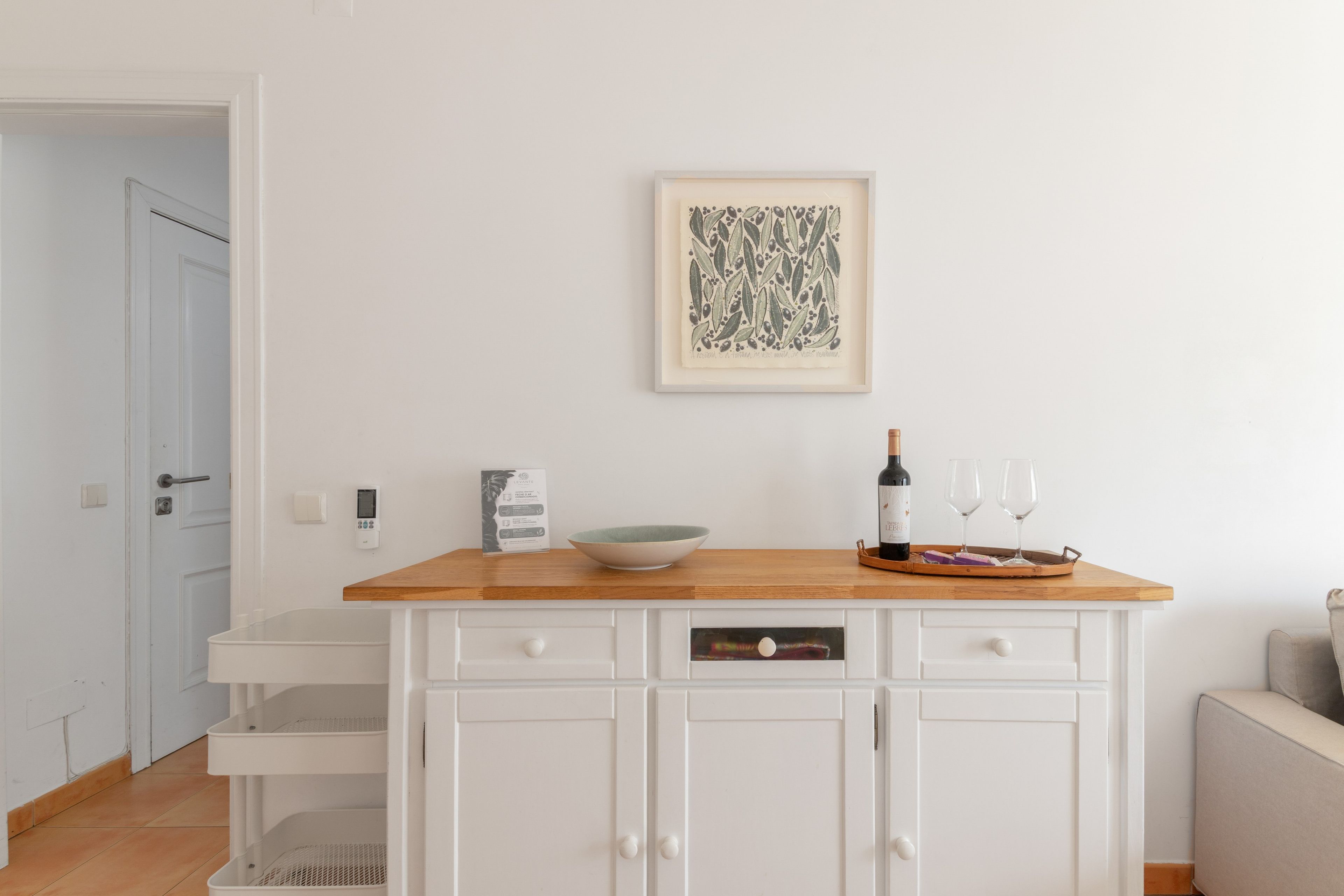 Een keuken met een houten aanrechtblad, een witte kast, een kunstwerk in een lijst, een wijnfles en twee glazen.