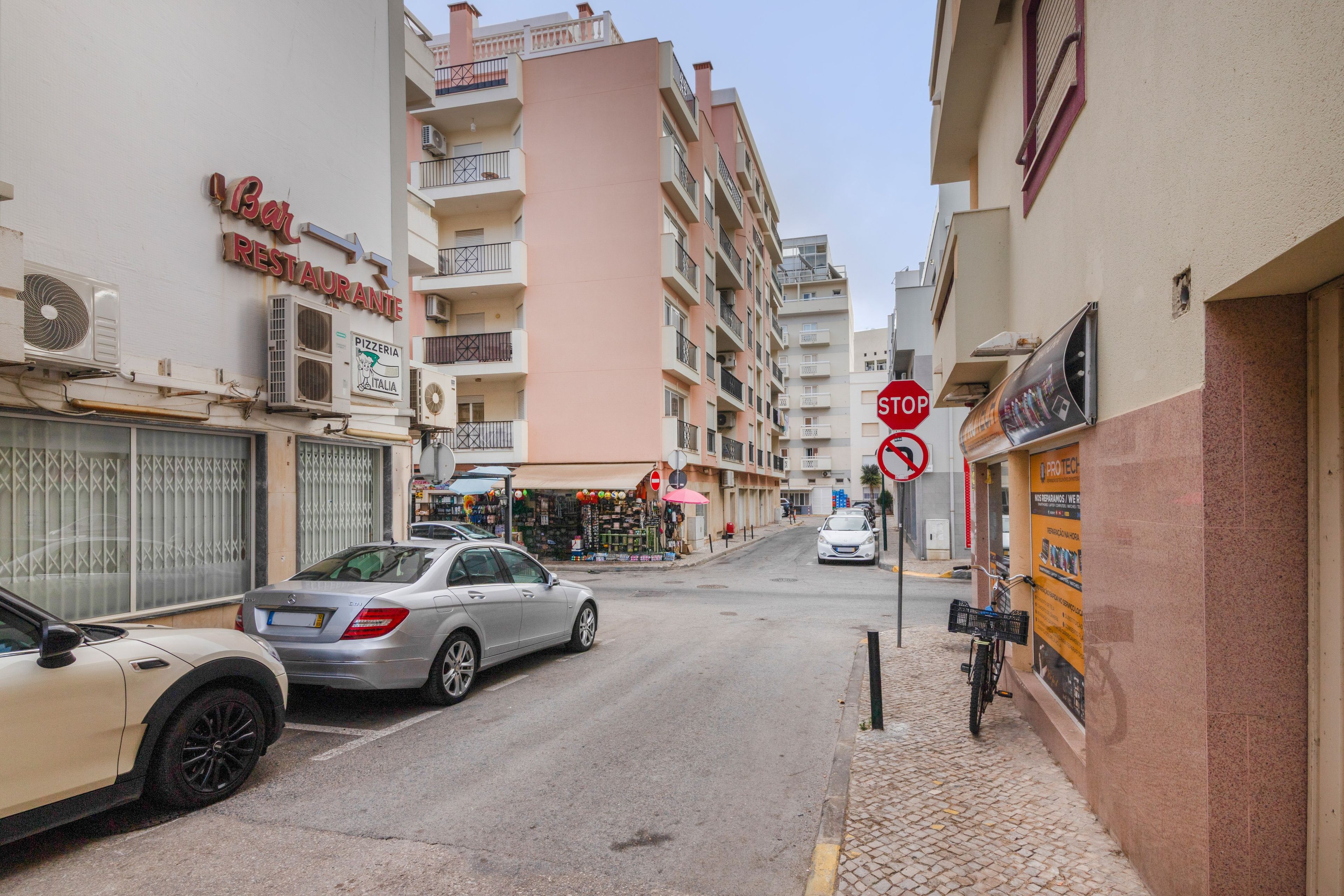 Une vue de rue avec des voitures garées, un panneau de signalisation indiquant un arrêt obligatoire et un bâtiment sur lequel on peut voir une enseigne de pizzeria.