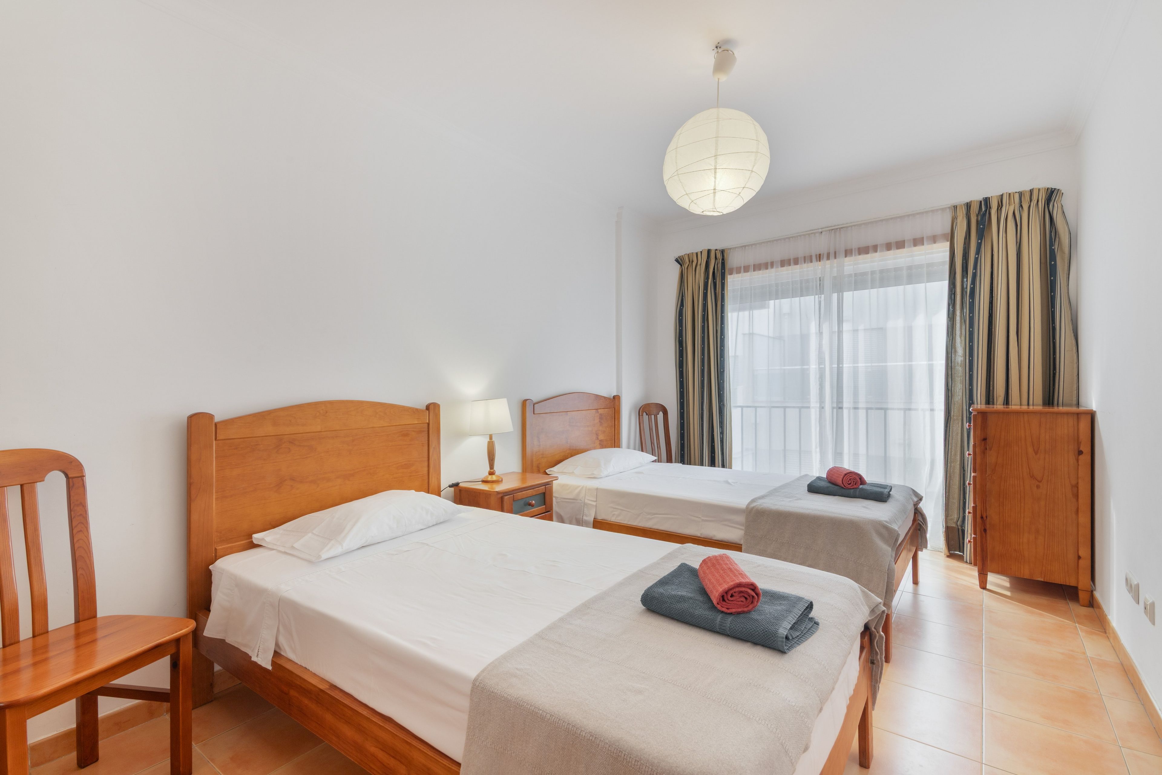 Habitación de hotel con dos camas, una silla de madera, una mesita de noche con lámpara, un armario y una ventana con cortinas.