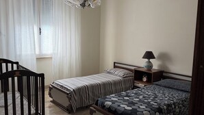 A bedroom with two beds, a crib, a chandelier, a bedside table with a lamp, and a window with sheer curtains.