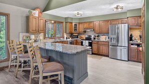 Une cuisine moderne avec des meubles en bois, un grand îlot central, des appareils électroménagers en acier inoxydable et une vue sur l’extérieur.