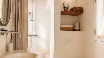 A bathroom with a freestanding sink, a toilet, a shower, and a wooden shelf with toiletries.