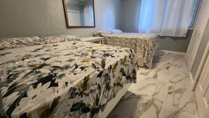 A hotel room with two beds, a small table, a mirror, an air conditioning unit, and a window with curtains.