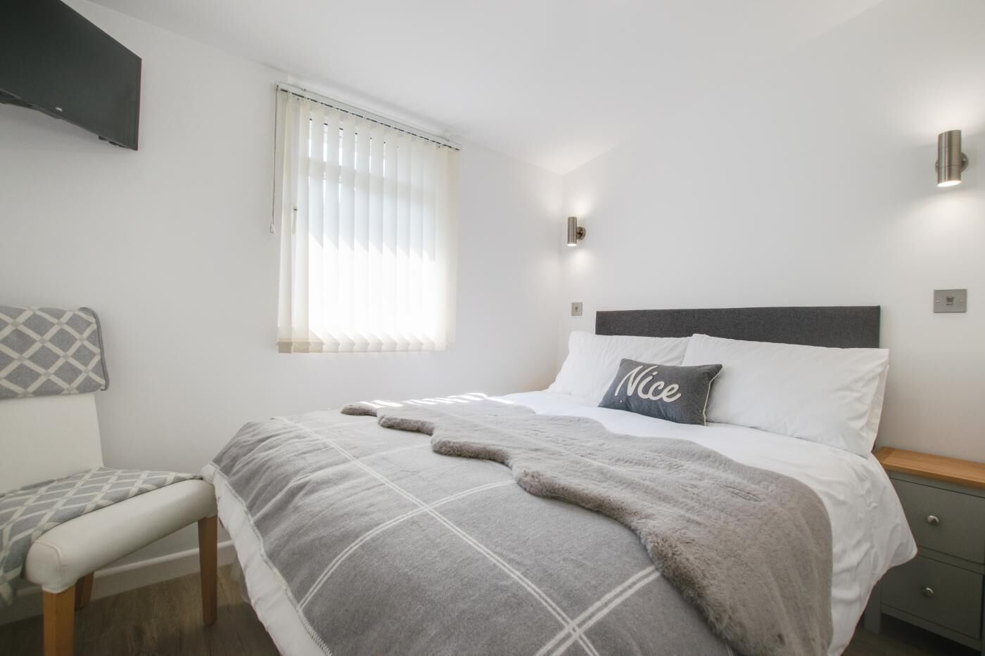 A bedroom with a bed, bedside table, chair, window with blinds, and wall-mounted light.