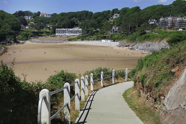A coastal walkway leading to a sandy beach with a view of a building and greenery in the background.