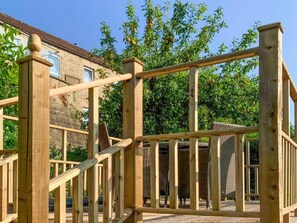 Una estructura de cubierta de madera con barandilla, un edificio de piedra al fondo y un árbol con hojas verdes.