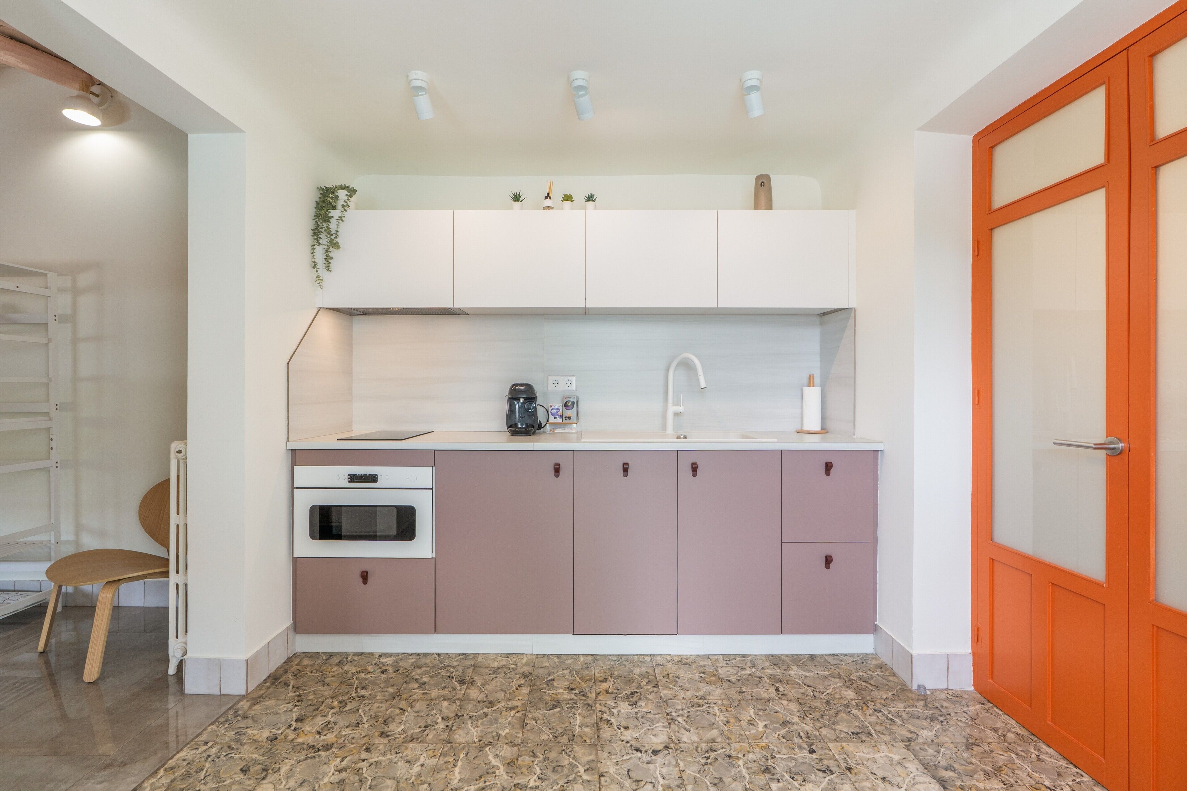 Una cocina moderna con gabinetes blancos y rosados, horno empotrado, cafetera y pileta.