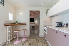 A compact kitchen with a countertop, two pink stools, a radiator, and a glimpse of a bedroom in the background.