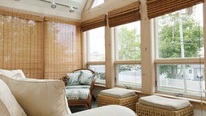 Une pièce comprenant un lit, un fauteuil en osier avec des coussins, une fenêtre donnant sur l’extérieur et un ventilateur de plafond.