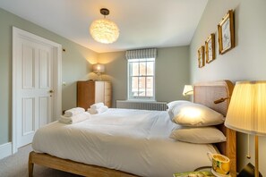 A neatly made bed with pillows and folded towels, a bedside lamp, a wooden cabinet, a radiator, a window with a view, and framed artwork on the wall. - 6 Bed in Southwold (oc-flgr) (Southwold)