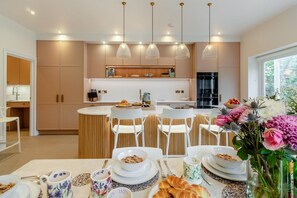 A modern kitchen with a central island, pendant lighting, and a dining area with a table set for breakfast. - 6 Bed in Southwold (oc-flgr) (Southwold)