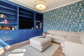 A living room with a blue entertainment unit, a beige sofa, and a patterned wallpaper. - 6 Bed in Southwold (oc-flgr) (Southwold)