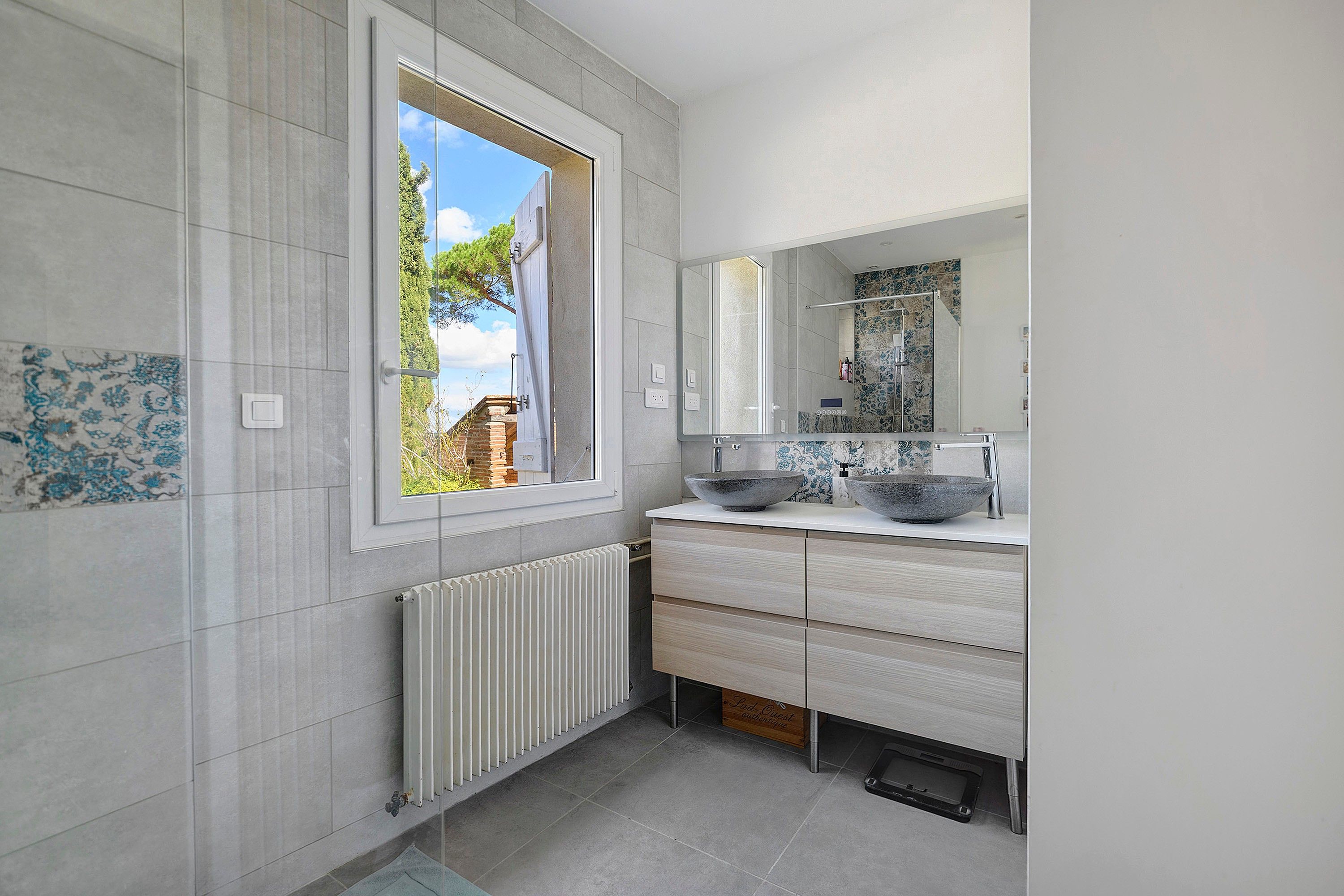 A modern bathroom with a large window, a white radiator, and a double sink with a bowl.