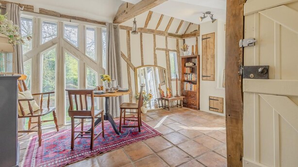 Een kamer met zichtbare houten balken, een groot raam, een tafel met stoelen, een boekenkast en een deur met slot.