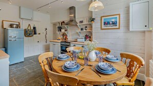 On aperçoit un coin repas avec une table en bois dressée pour quatre personnes, entourée de chaises en bois. Au fond, on distingue une cuisine avec un réfrigérateur bleu et des placards blancs.