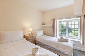 A hotel room with two beds, a nightstand with a lamp, a shelf above the bed, and a window with curtains. - 3 Bed in Pancrasweek (oc-t28012) (Pancrasweek)