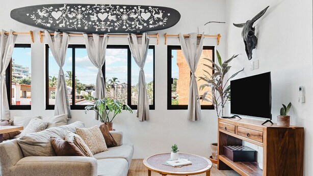 Ein Wohnzimmer mit einer Couch, Fernseher, Holzschrank und großen Fenstern mit Vorhängen.
