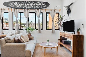 Ein Wohnzimmer mit einer Couch, Fernseher, Holzschrank und großen Fenstern mit Vorhängen.