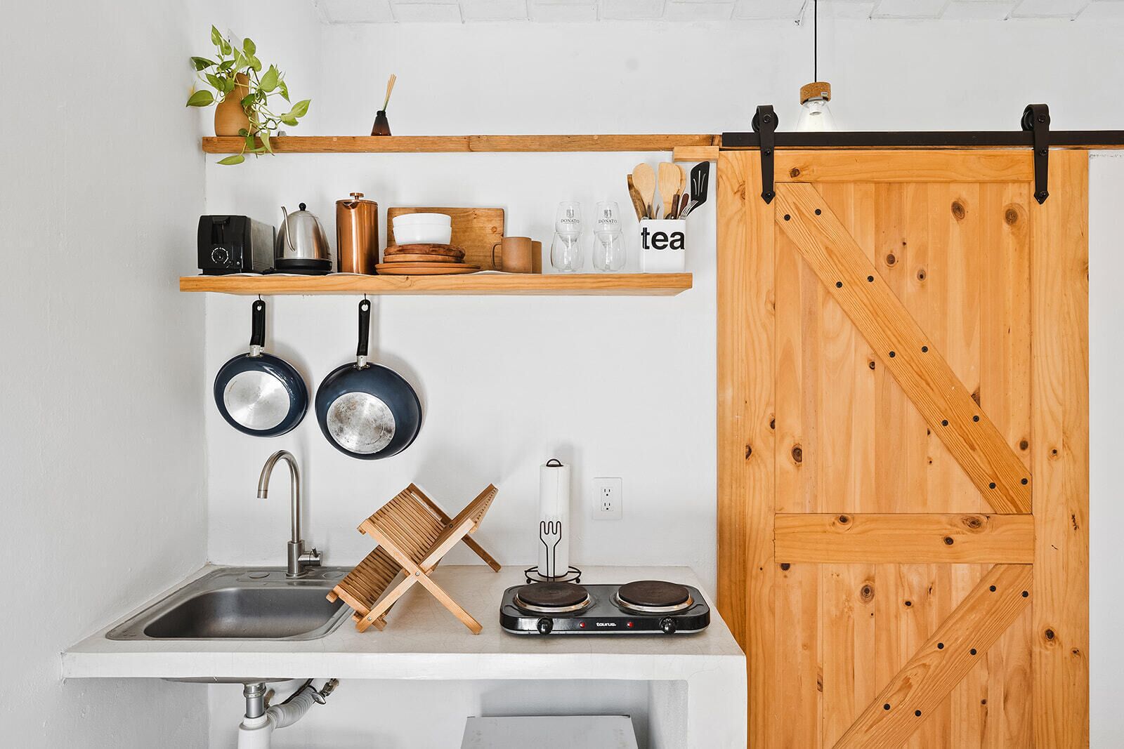 Een keuken met een spoelbak, twee koekenpannen, een waterkoker, een koffiezetapparaat, een theeset en een houten schuifdeur.