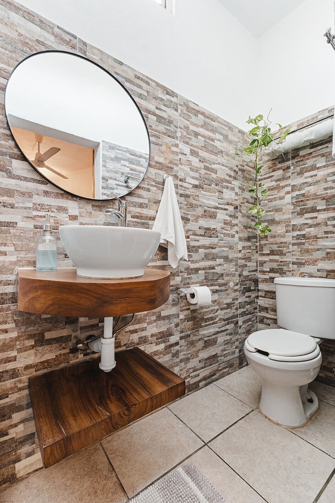 A bathroom with a round mirror, a wooden vanity, a white sink, a toilet, and a patterned wall.