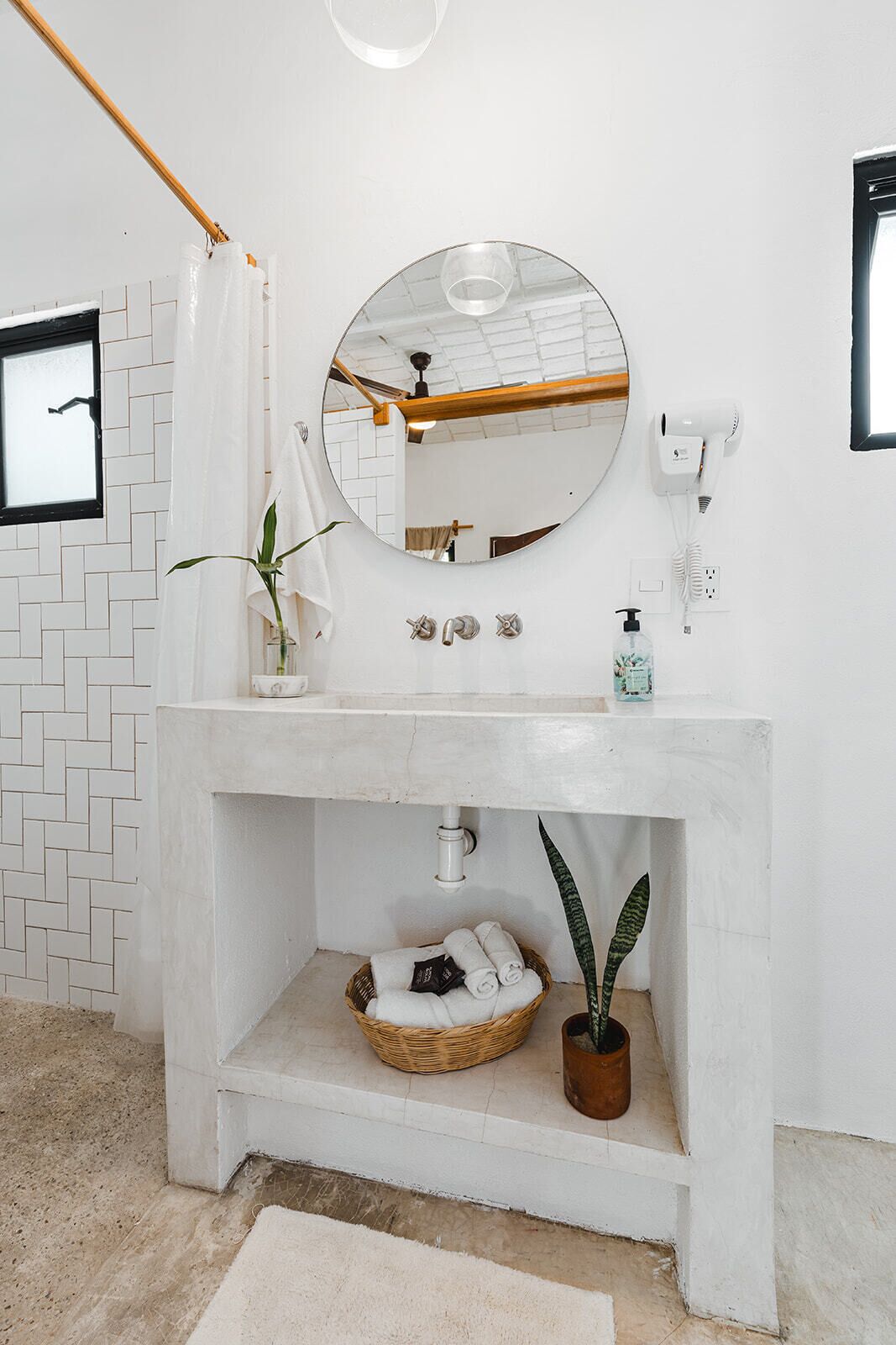 Un baño con lavamanos blanco, espejo, cortina de ducha, toallero, planta y cesta con toallas.