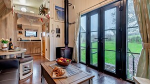 A cozy interior with a large black stove, a dining area with a table and chairs, a kitchen with wooden cabinets, and a view of a green landscape through the glass doors. - 2 Bed in Ash (oc-o29006) (Stoke Fleming)