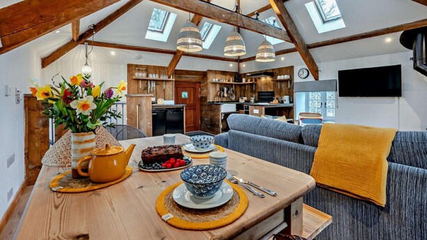 Een knus loft-appartement met houten plafond, zichtbare balken en dakramen. Een eethoek met een tafel gedekt voor twee personen, een bank en een aan de muur bevestigde televisie.
