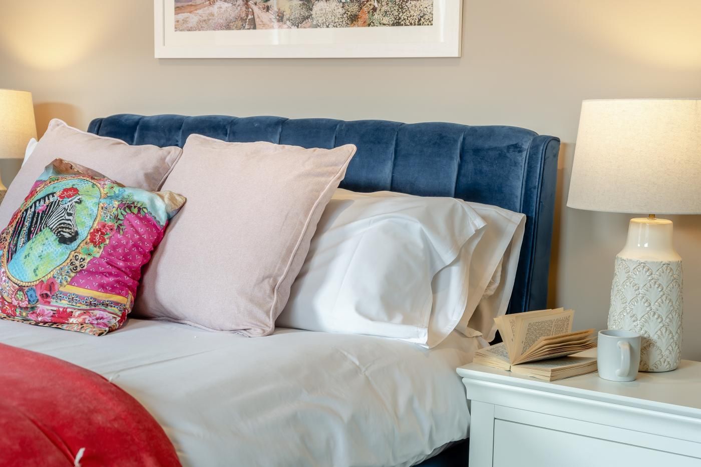 Un lit bien fait, avec une tête de lit bleue, un coussin coloré et un coussin blanc. On y trouve une table de chevet blanche, sur laquelle sont posés une lampe et un livre.