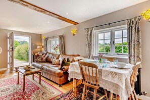 Ein Wohnzimmer mit einer Couch, einem Esstisch für zwei Personen und einem großen Fenster mit Vorhängen.
