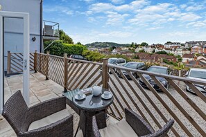 Un balcon agrémenté de meubles en osier, d’une table et offrant une vue sur un quartier résidentiel où sont garées des voitures.