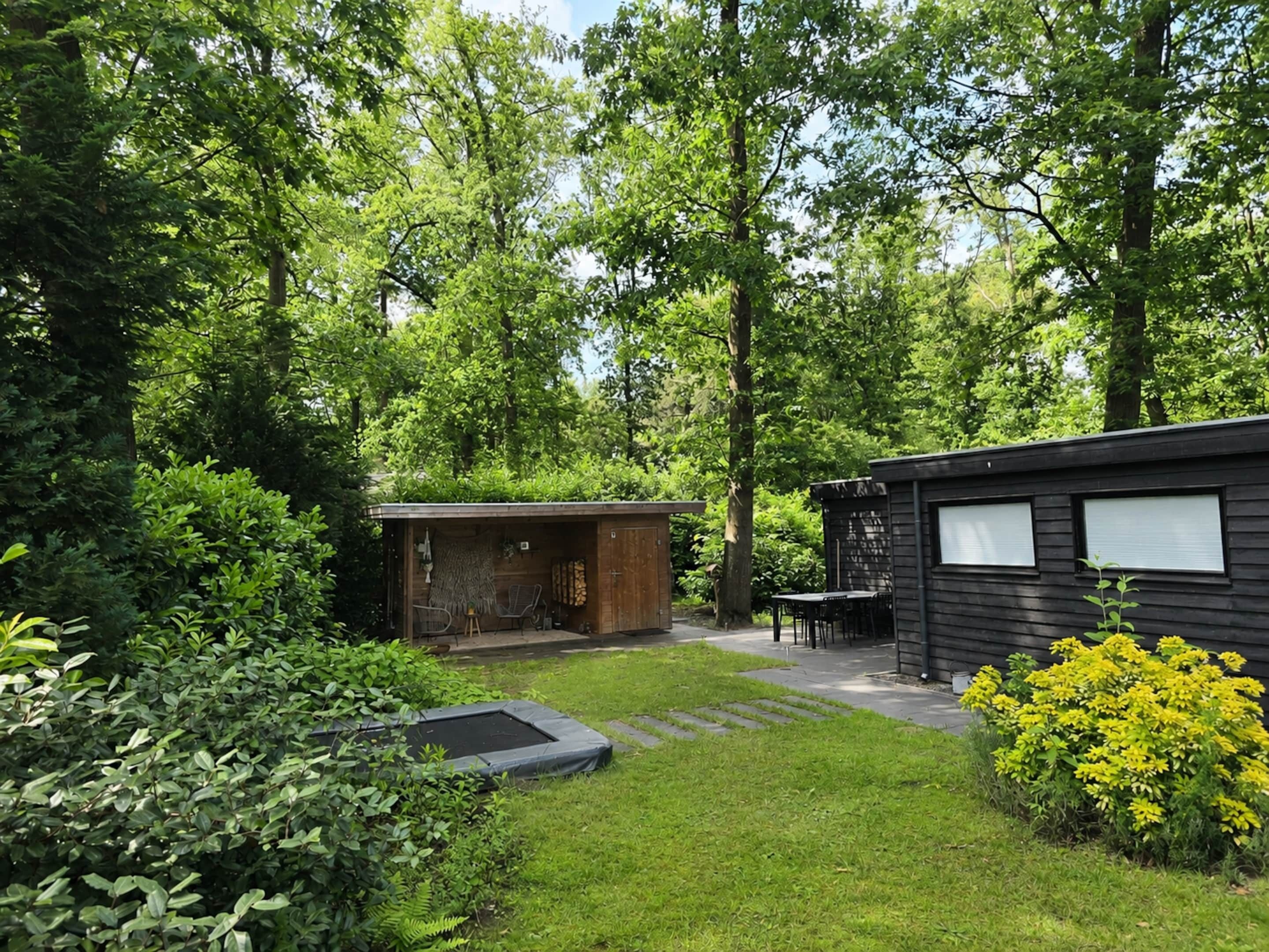 Eine kleine Holzhütte mit einer Holzterrasse und einem Trampolin in einem üppigen, grünen Garten.