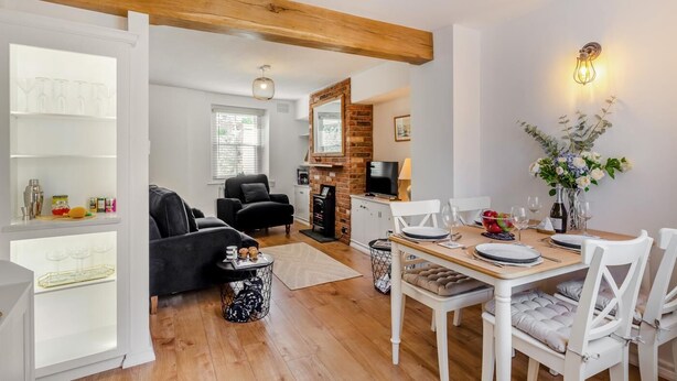 A modern living room with a fireplace, a dining area with white chairs, and a kitchen in the background. - 2 Bed in Appledore (oc-m29918) (Appledore)