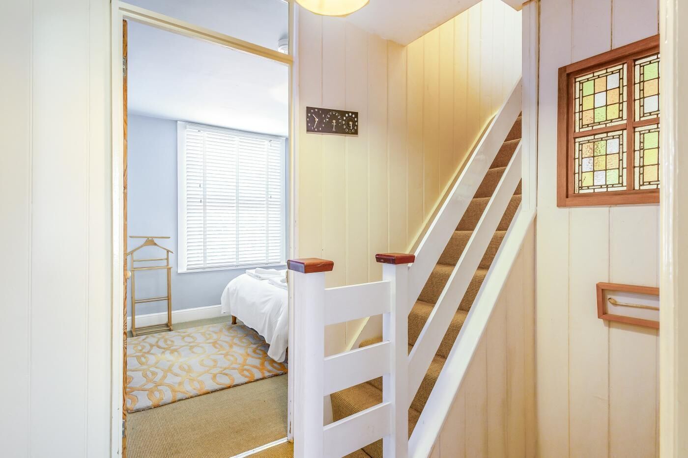 Un escalier menant à une chambre à coucher équipée d’un lit, d’une fenêtre avec des stores et d’une horloge murale.