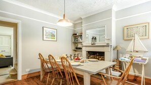 Et spiseområde med et bord dækket til fire personer, træstole, en hængende lampe, en pejs og en bogreol.