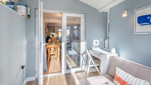 Huoneessa on liukuovi, joka johtaa ruokailutilaan, valkoinen työpöytä, tyynyillä varustettu sohva ja seinälle asennettu valaisin.