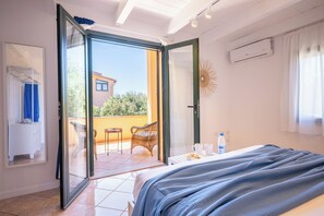 Une chambre à coucher avec un lit, une petite table sur laquelle se trouve une bouteille, une chaise et un balcon d’où l’on peut admirer un immeuble et des arbres.