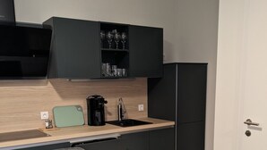 Une cuisine moderne avec un plan de travail en bois, un évier, une machine à café et un meuble de rangement au-dessus du plan de travail. On y trouve une table à manger avec des chaises et une porte sur le côté droit.