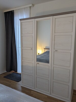 A bedroom with a large wardrobe, a bed, a bedside table, and a window with curtains.