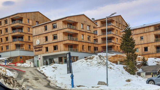 Un bâtiment en bois à plusieurs étages, doté de balcons, avec une enseigne indiquant « Almresort Nassfeld », et un sol recouvert de neige.