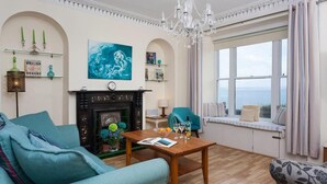 Een kamer met een open haard, uitzicht op zee, een kroonluchter en een vensterbank met kussens.