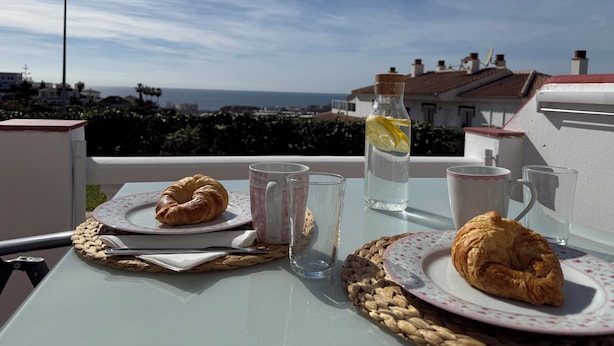 Une table avec des croissants, une bouteille d’eau et une tasse, donnant sur une vue sur la côte.