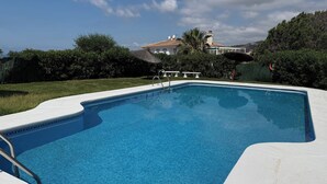Une piscine à bord incurvé, entourée d’une terrasse blanche, et une maison avec un toit en tuiles en arrière-plan.
