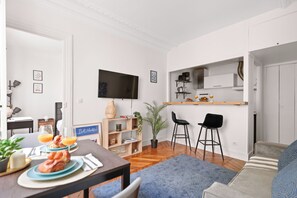 A modern living room with a dining area, a flat-screen TV mounted on the wall, a kitchen area with a wooden countertop, and a blue sofa. - Cozy 1BR Near Eiffel Tower - Sleeps 4 (Paris)