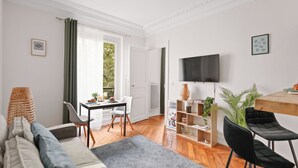 Ein modernes Wohnzimmer mit einer Couch, einem Essbereich mit Tisch und Stühlen, einem an der Wand montierten Fernseher und einem Bücherregal mit Pflanzen.