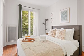 Une chambre à coucher avec un lit, une fenêtre avec des rideaux, un radiateur, une table de chevet avec une lampe et des œuvres d’art accrochées au mur.