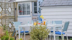 Ein weißes Haus mit blauer Tür, ein weißer Tisch mit blauen Stühlen und ein Garten davor.