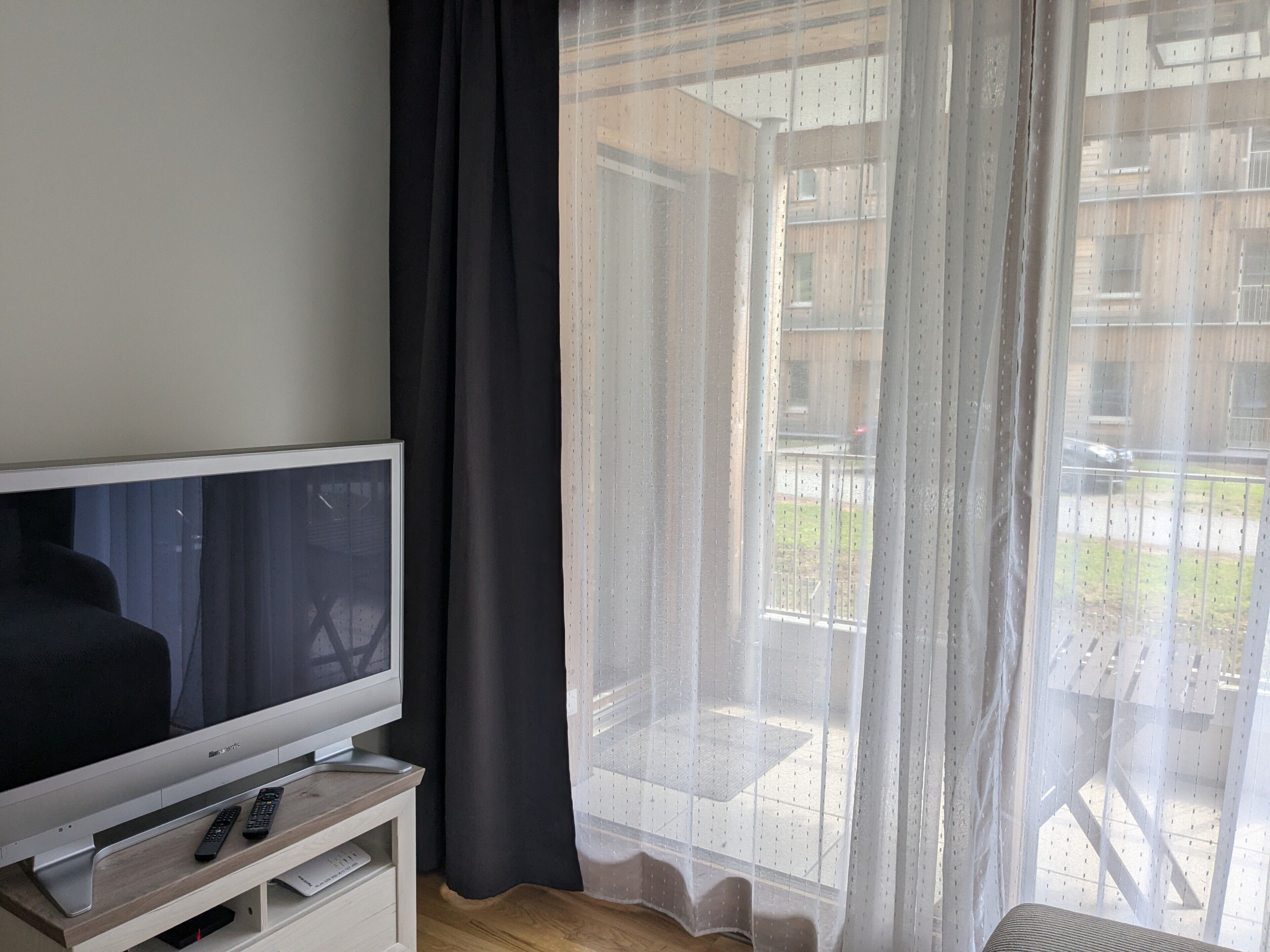 A room with a flat-screen TV on a wooden stand, a remote control, and a sheer curtain partially drawn.