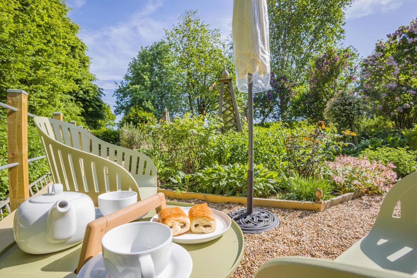 A garden setting with outdoor seating, a table with a teapot, cups, and pastries, surrounded by lush greenery and flowers.