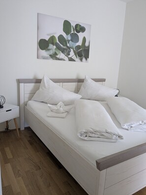 Un lit bien fait, avec du linge de lit blanc et des oreillers, une table de chevet avec une lampe, et un tableau représentant des feuilles d’eucalyptus accroché au mur.
