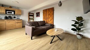 A modern living space with a sofa, a round wooden coffee table, a potted plant, a kitchen area with wooden cabinets, and a microwave. - Neues Appartement im Lepels Hof (Lütow)